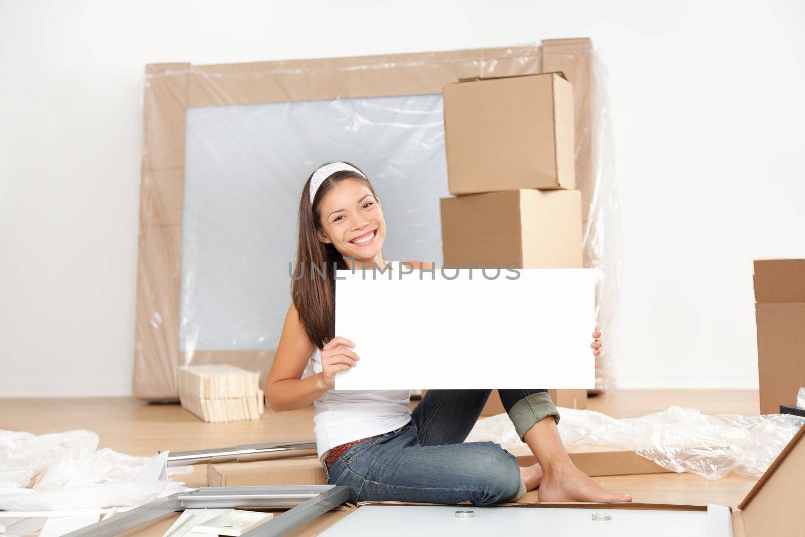 Moving sign woman. Woman moving in new home unpacking showing blank white sign while doing furniture assembly of new table. Beautiful young mixed race Asian Caucasian young woman in new apartment condo.