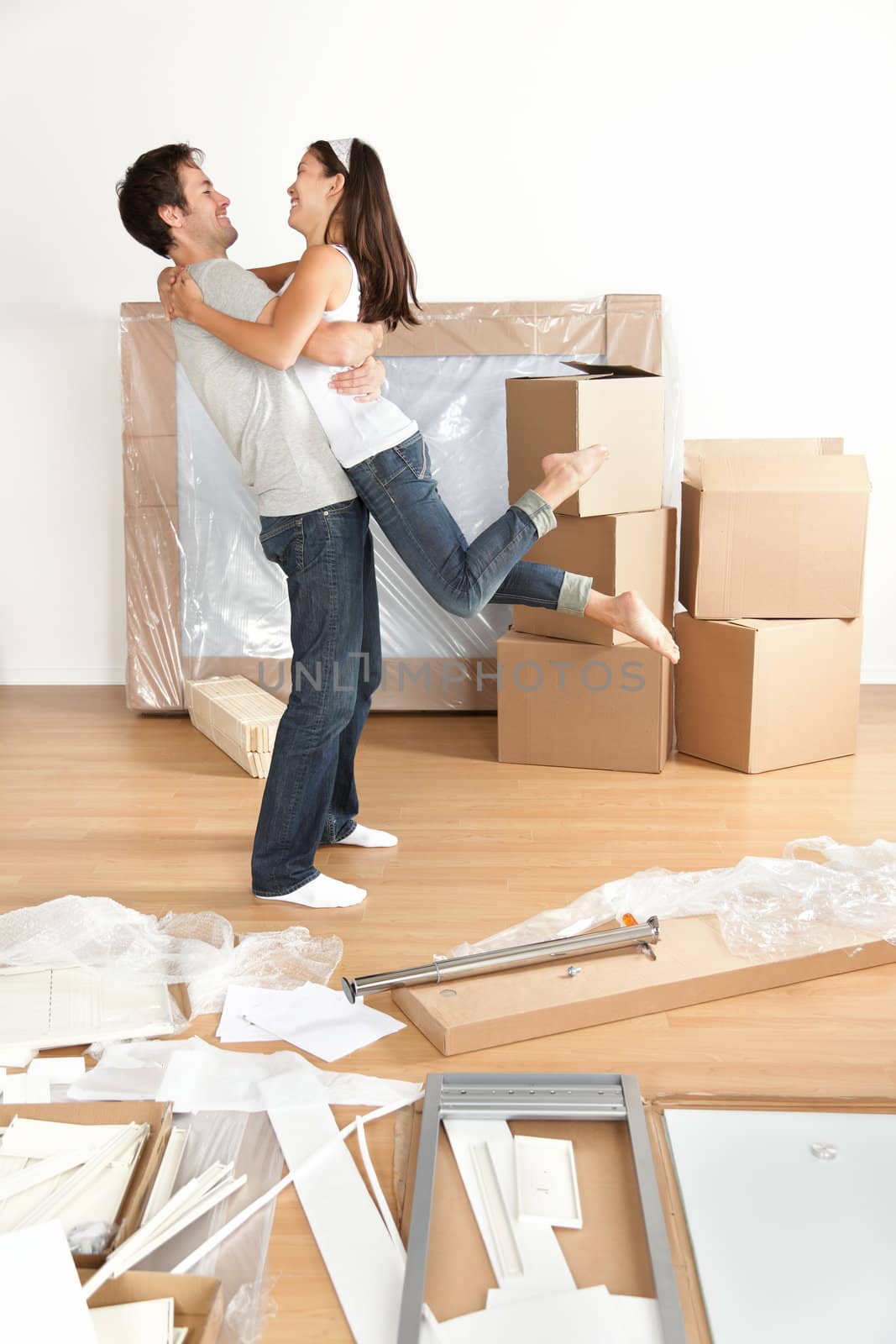 Couple moving in happy and excited in new home. Young interracial couple with moving boxes and furniture assembly in new house or apartment. Caucasian man and Asian woman embracing.