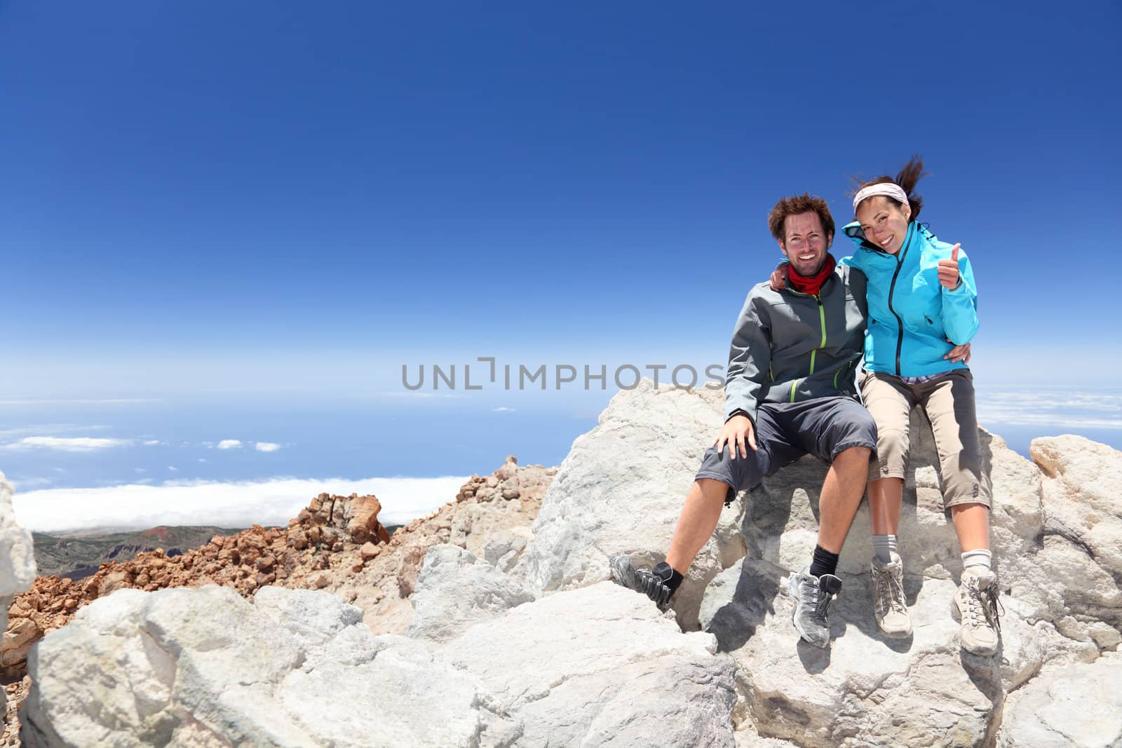 Couple on mountain top of volcano Mount Teide, Tenerife after hiking to summit. Woman showing thumbs up success sign.