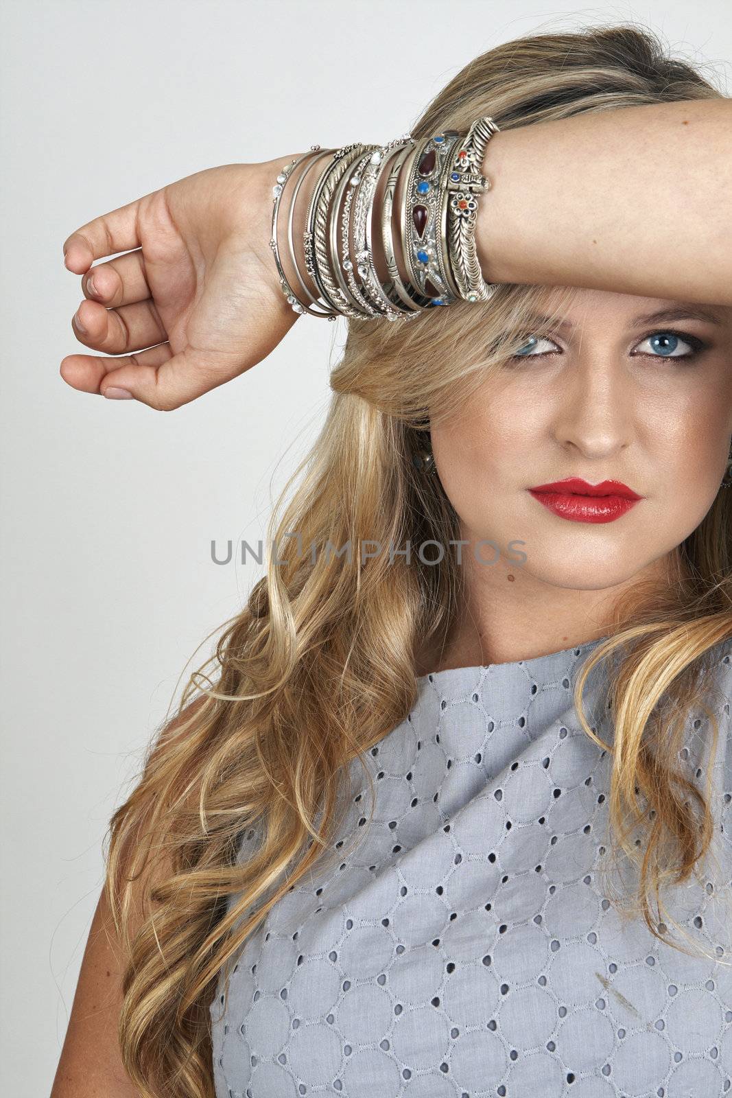 Portrait of a beautiful blonde woman with light blue eyes and classic make-up with lots of bangles on her arm on grey background