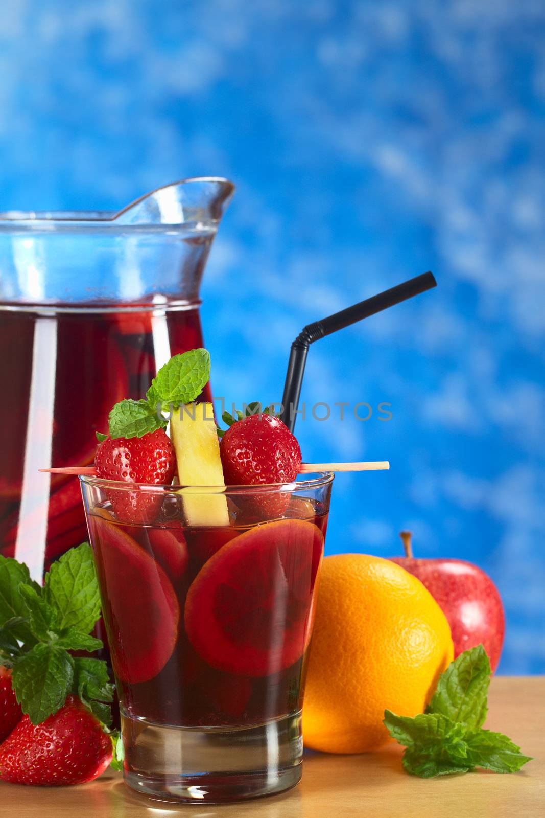 Refreshing red wine punch called sangria mixed with orange, apple and mango, garnished with strawberries and pineapple on skewer with a jug of sangria in the back and fruits around (Selective Focus, Focus on the fruits on the skewer)