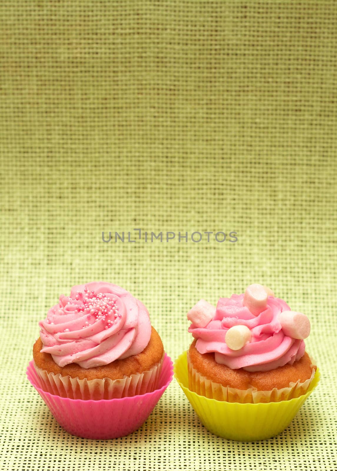 Two fresh vanilla cupcakes in pink and green cups with strawberry icing and marshmallows on hessian background