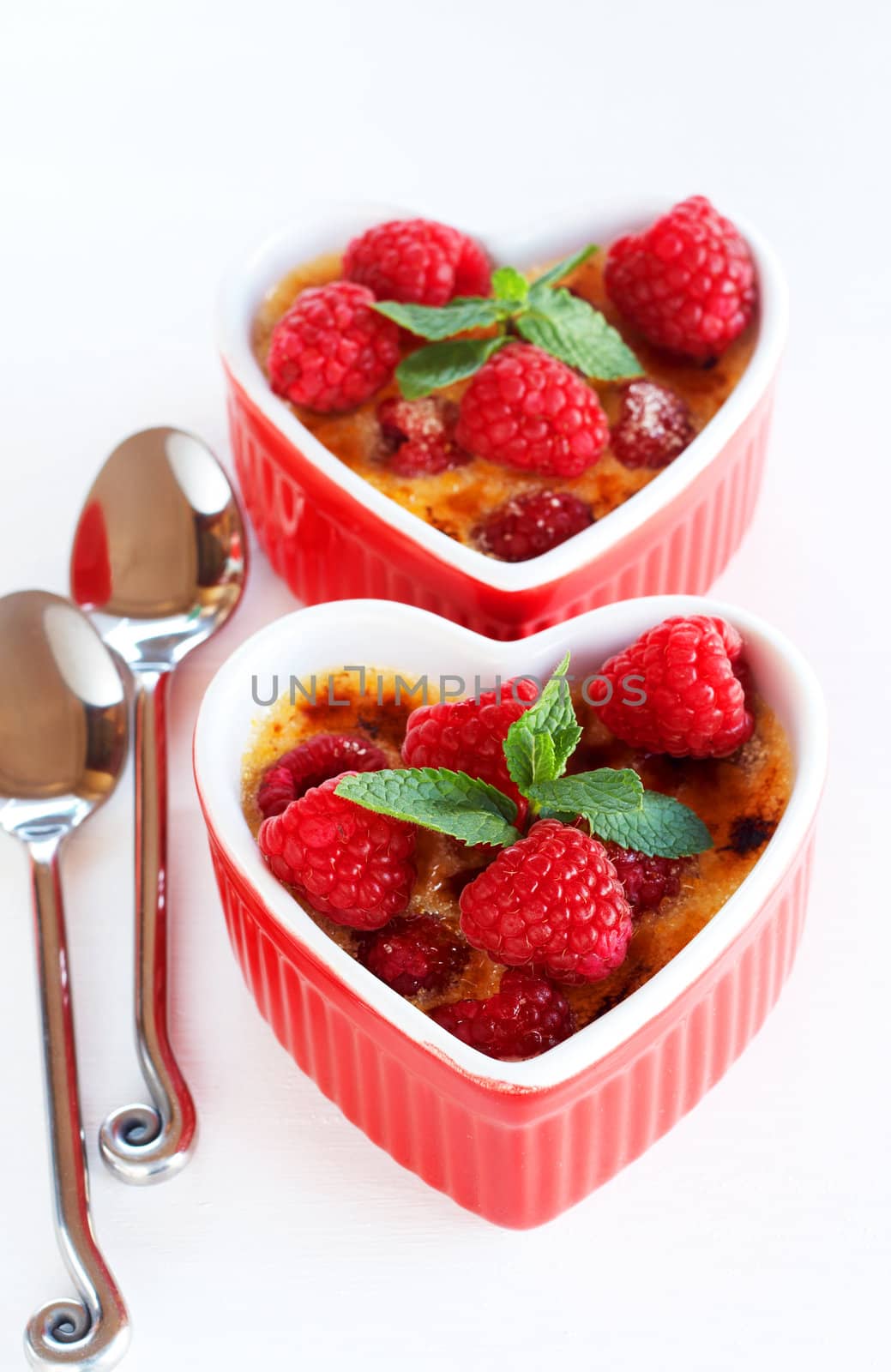 French creme brulee dessert with raspberries and mint covered with caramelized sugar in red heart shaped ramekins on white background