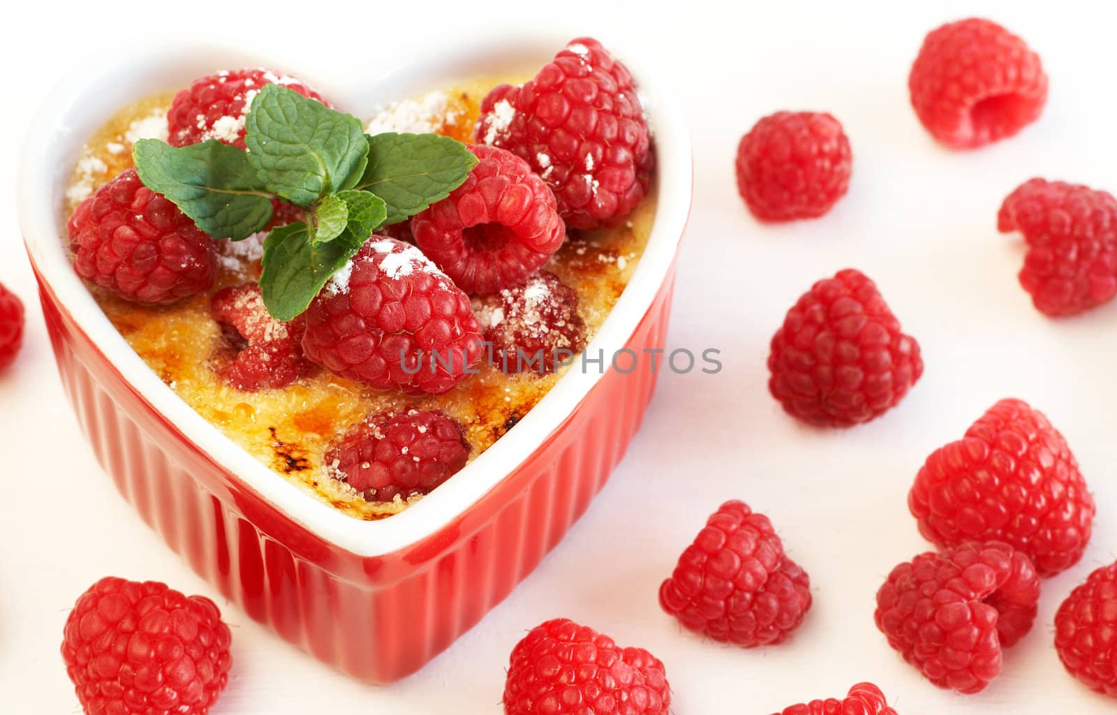 French creme brulee dessert with raspberries and mint covered with caramelized sugar in red terracotta heart ramekin on white background