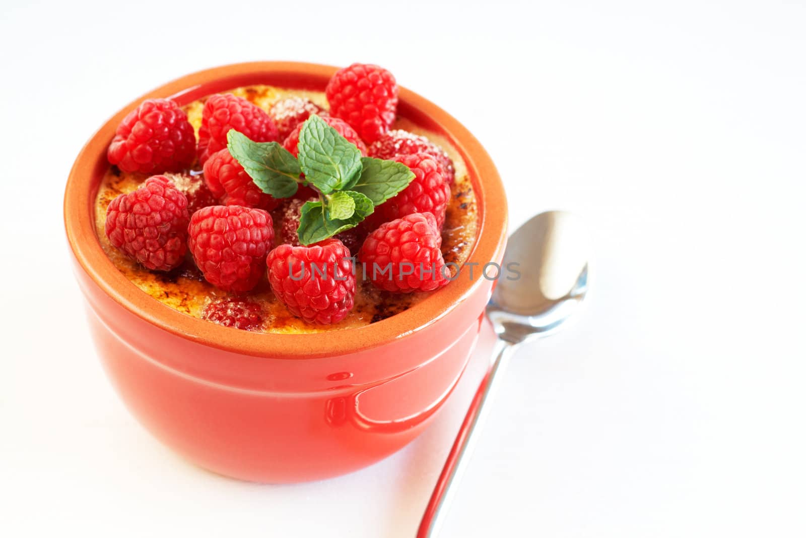 French creme brulee dessert with raspberries and mint covered with caramelized sugar in red terracotta ramekin on white background