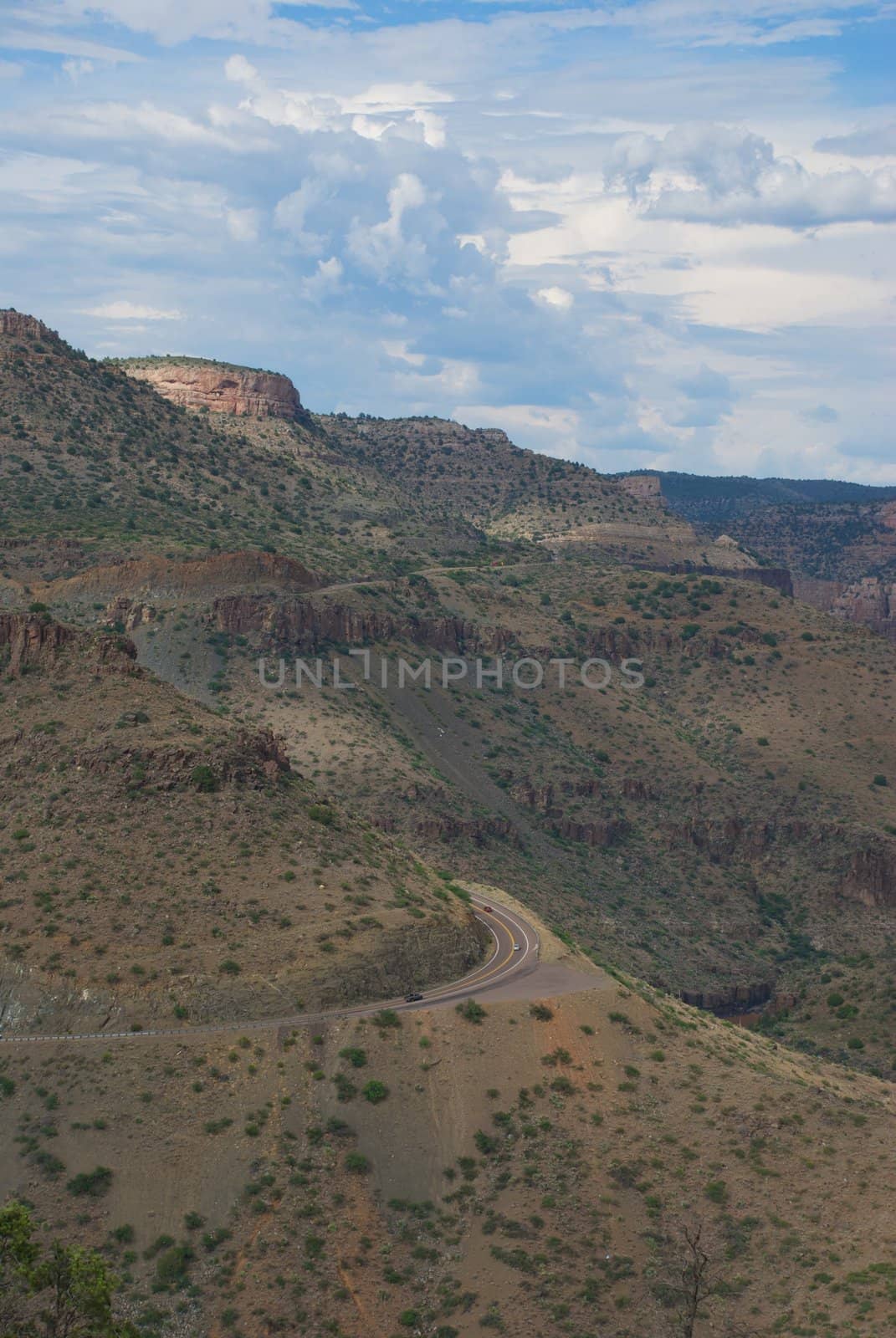 Huge desert cliffs loom over a winding highway road with swirling white clouds and a hint of blue sky