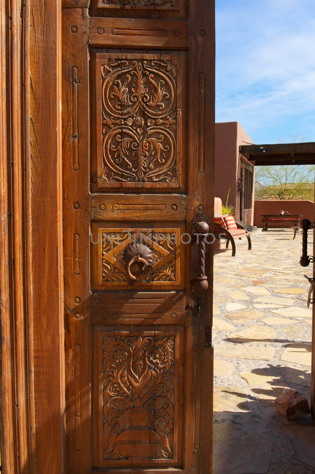 A wooden, ornate, opened door with metal knocker leads to a inner courtyard with stone and seating