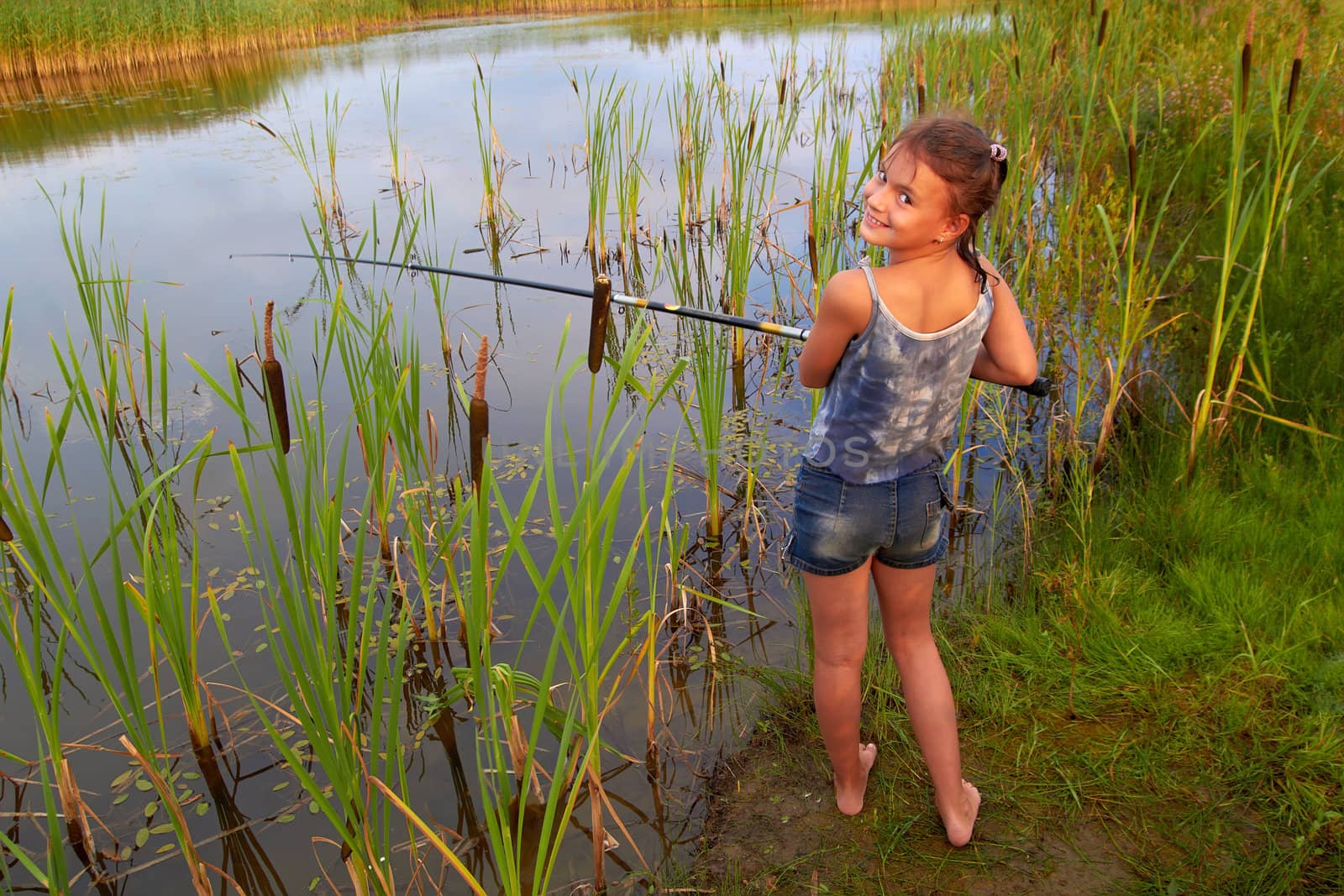 pretty young smiling girl fishing with long rod  on river outside in summer sunshine