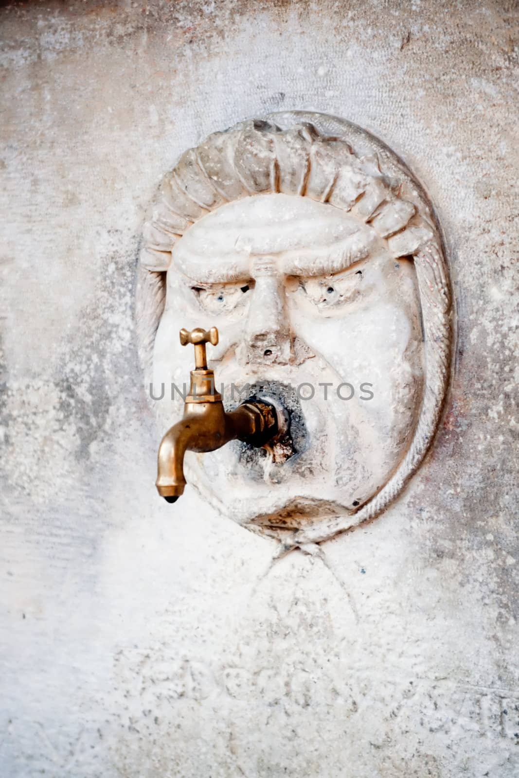 Public water-tap with historic stone carved face on marble wall.