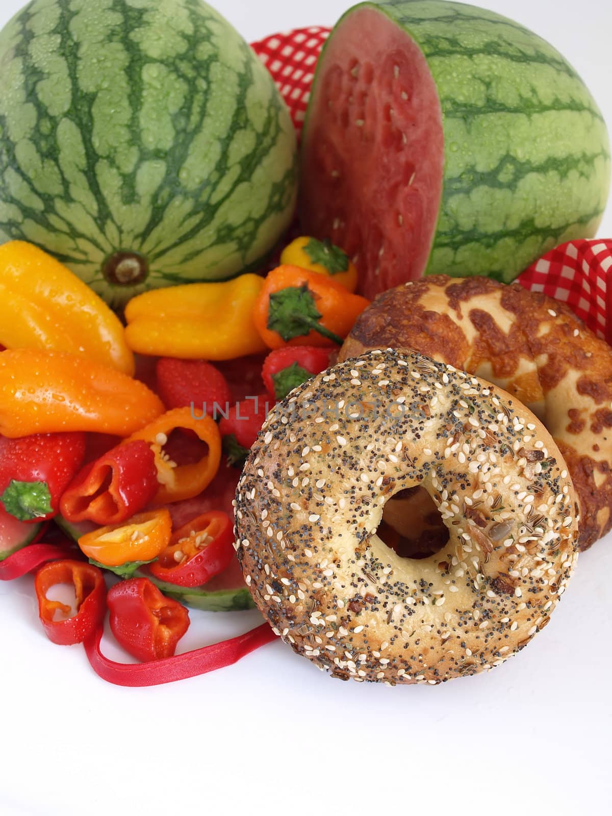 Beautiful red, orange and yellow peppers, ripe watermelon, and bagels set against a white background.