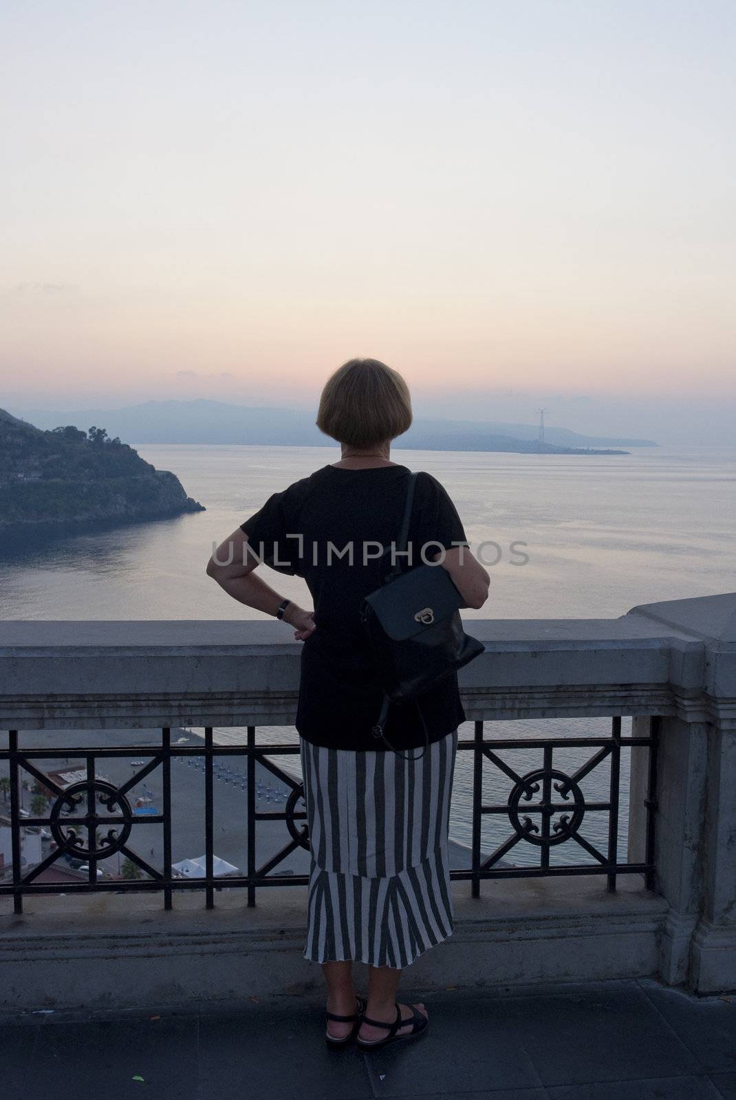 Senior female tourist at sunset in Scilla - the region of Calabria, Italy. She is looking at the strait of Messina with Sicily in the background.