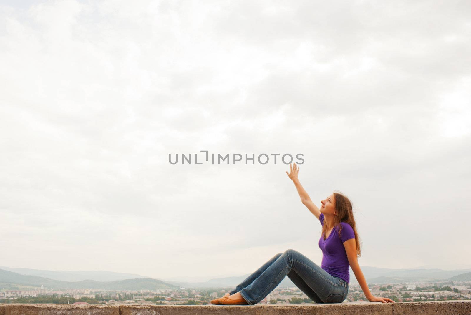 Young woman sitting with raised hand against blue sky