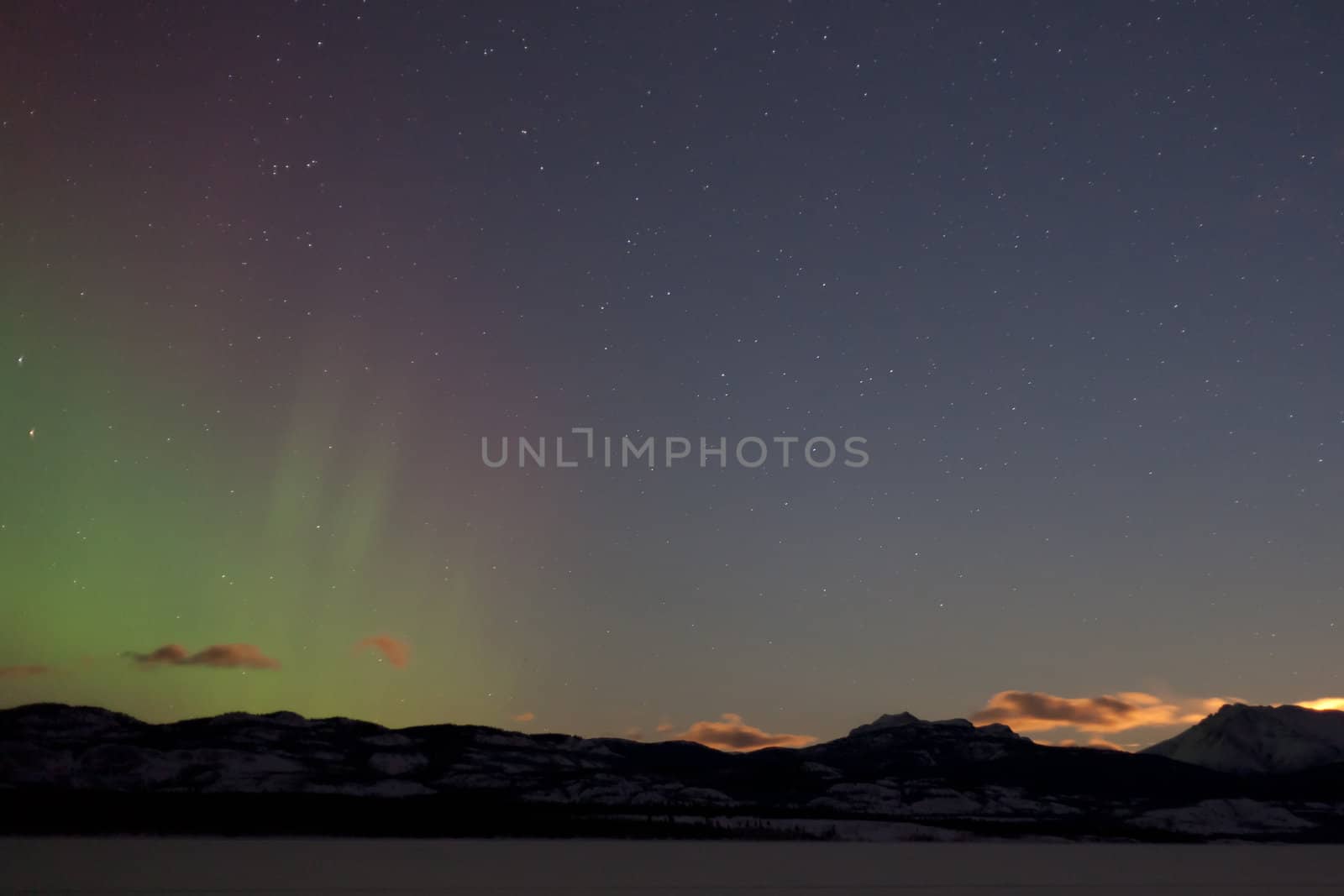Northern lights (aurora borealis) and rising moon above and behind snowy mountain range in winter.