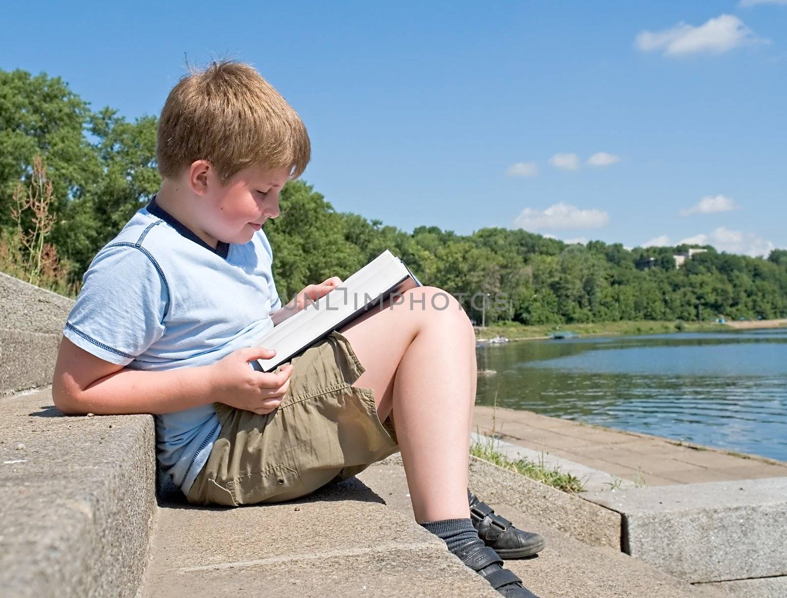 A little boy reads a big book with river at background