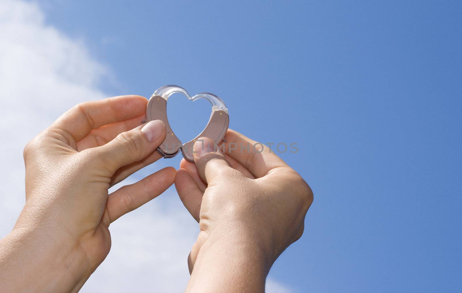 Hands showing a heart shape from digital hearing aids in fron of a blue sky background