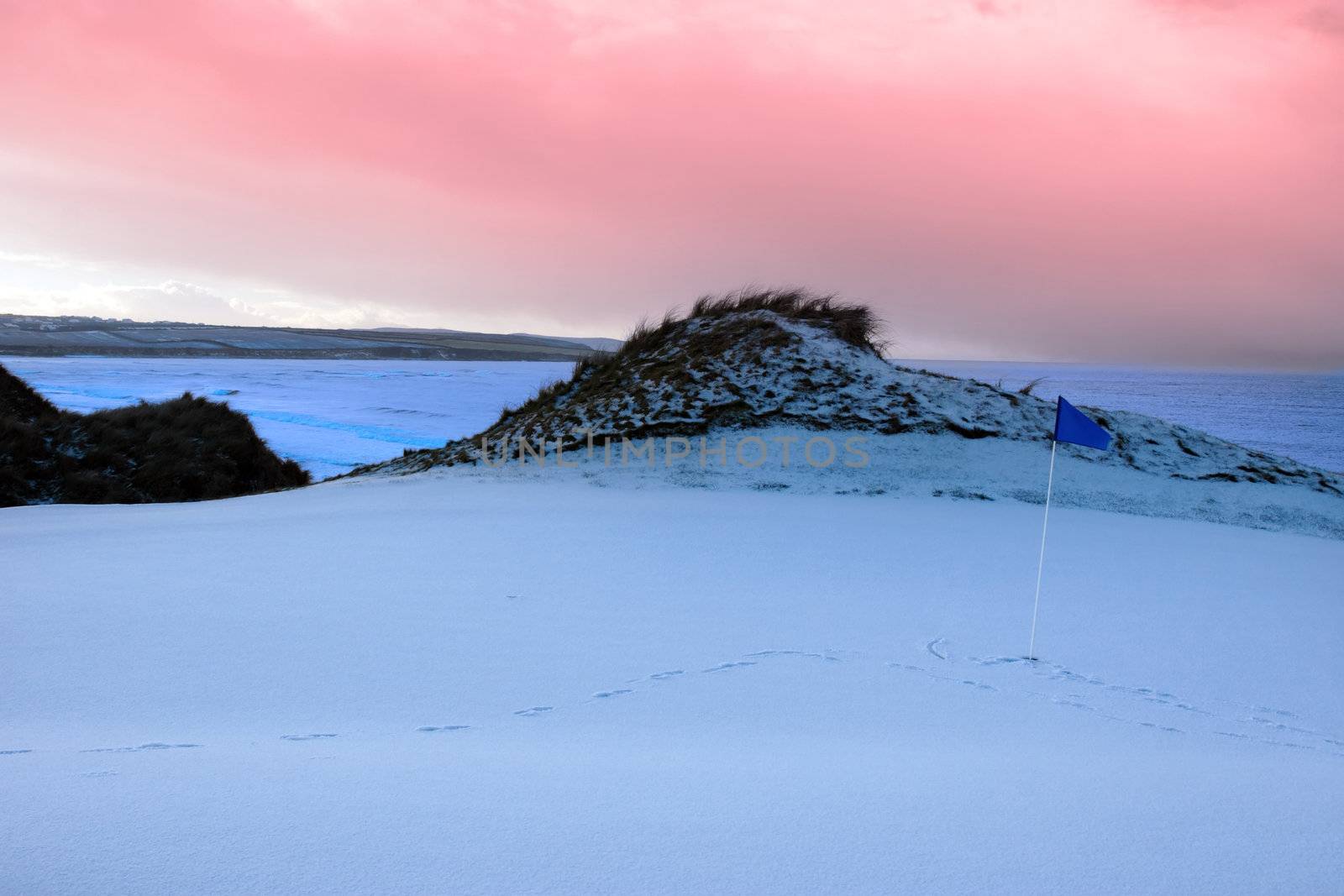 a snow covered links golf hole in ireland with blue flag and red sunset sky