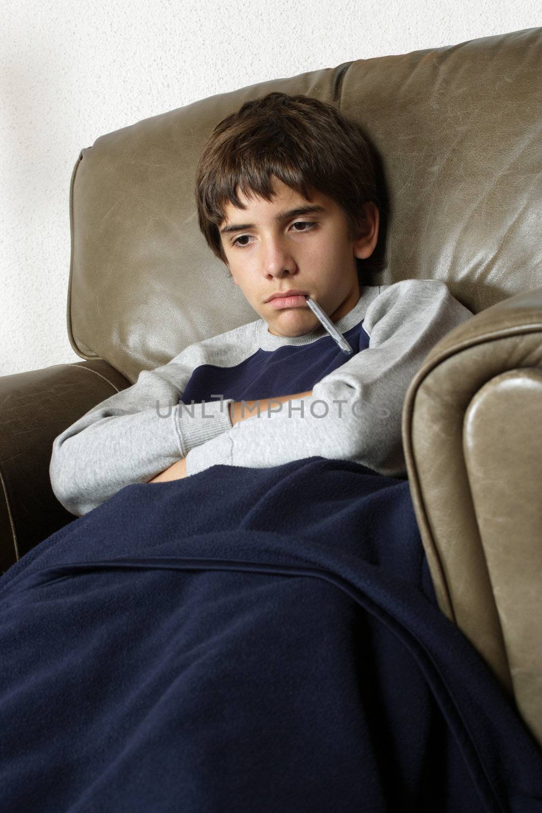 Sick child sitting in big leather chair with a thermometer in his mouth.
