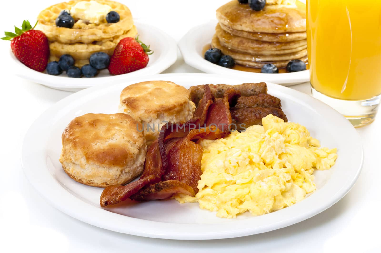 Breakfast plate with scrambled eggs, bacon, and buttermilk biscuits.  Waffles, pancakes, and orange juice in background.  Isolated on white background.