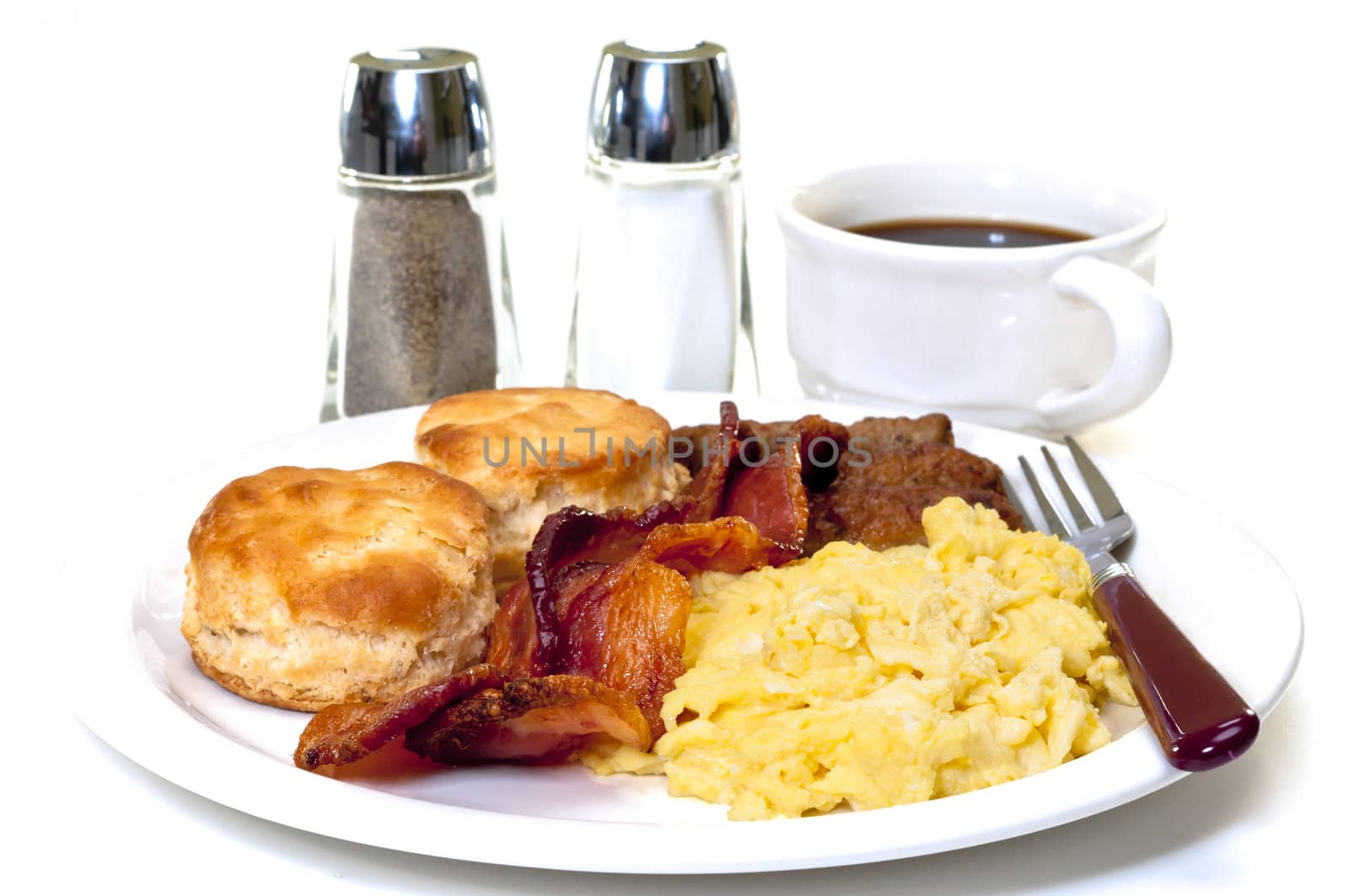 Big country breakfast with scrambled eggs, bacon, sausage, buttermilk biscuits, and coffee.  Salt and pepper shakers in background.  Isolated on white background.