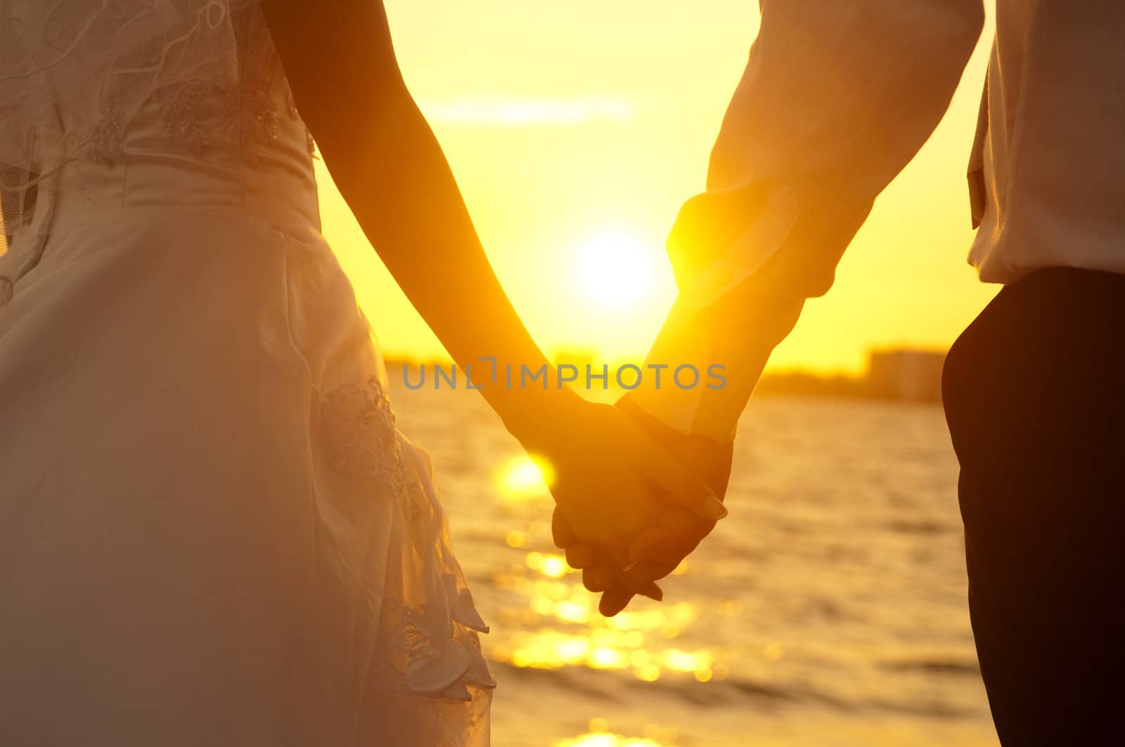 Young adult male groom and female bride holding hands on beach at sunset.