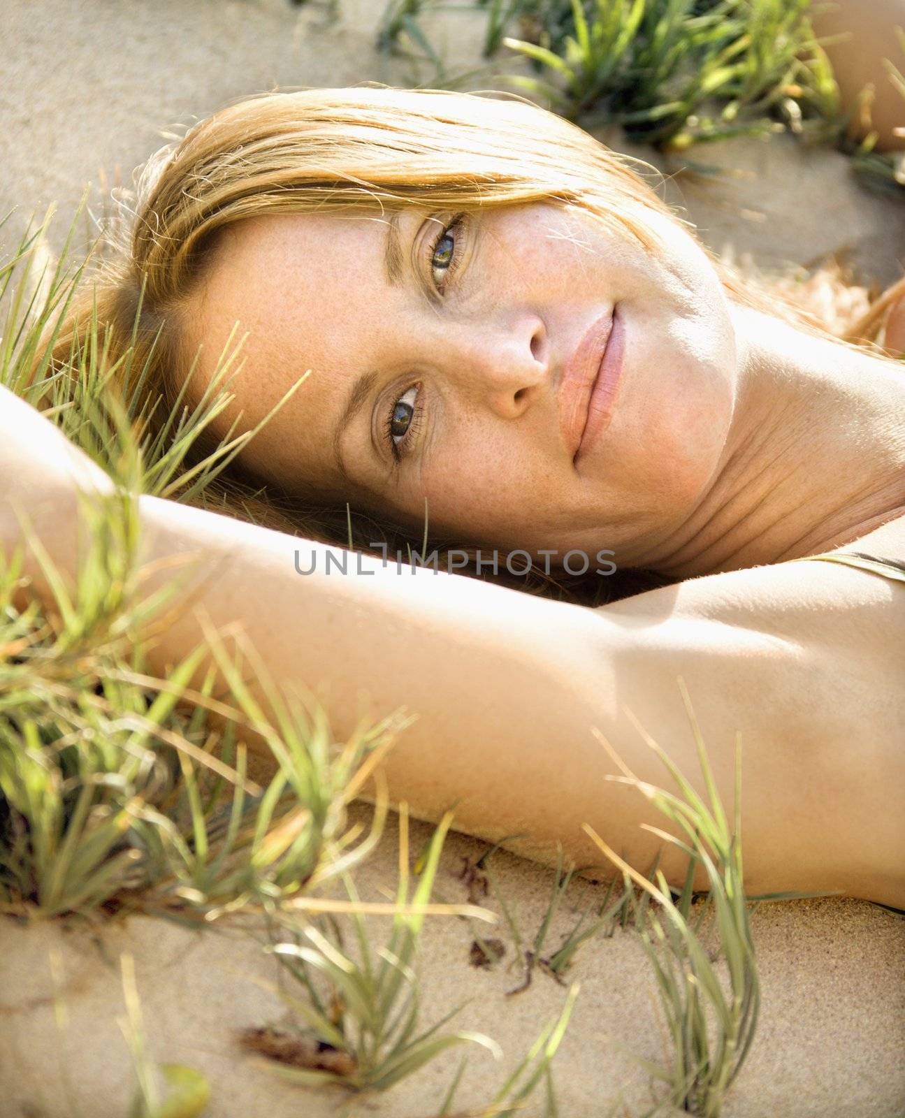 Close up portrait of attractive redheaded woman lying in grassy sand looking at viewer.