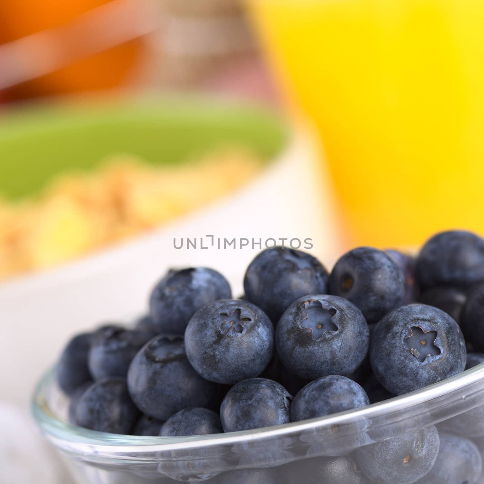 Fresh blueberries in glass bowl with a bowl of cereal and orange juice in the back (Selective Focus, Focus on the middle row of blueberries)