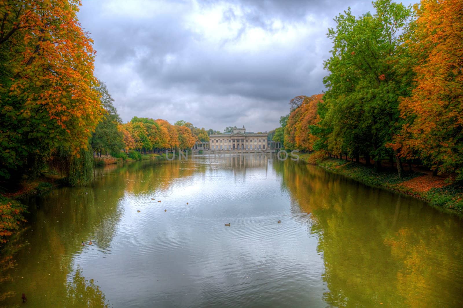 lazienki park warsaw poland in autumn, showing trees of different colors, lake or pond reflecting a historic building, clouds