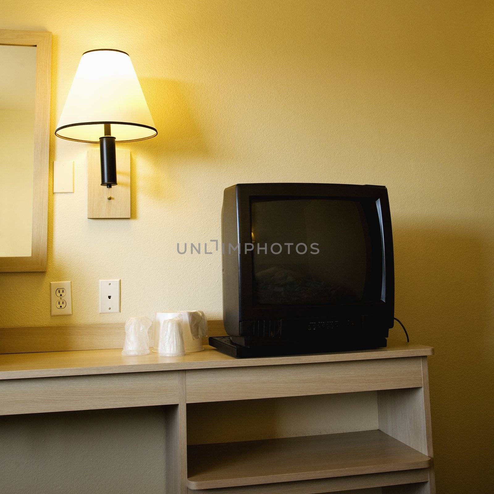 Interior shot of motel room with TV set on nightstand next to wall lamp.