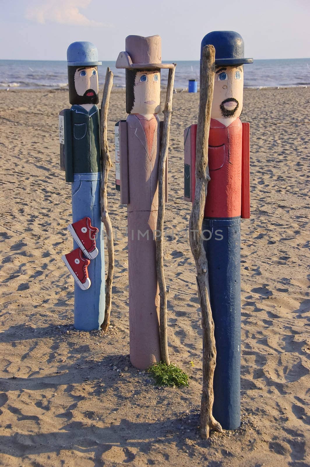 Painted wood carvings of two bearded men and a woman in pink with hats lined on beach