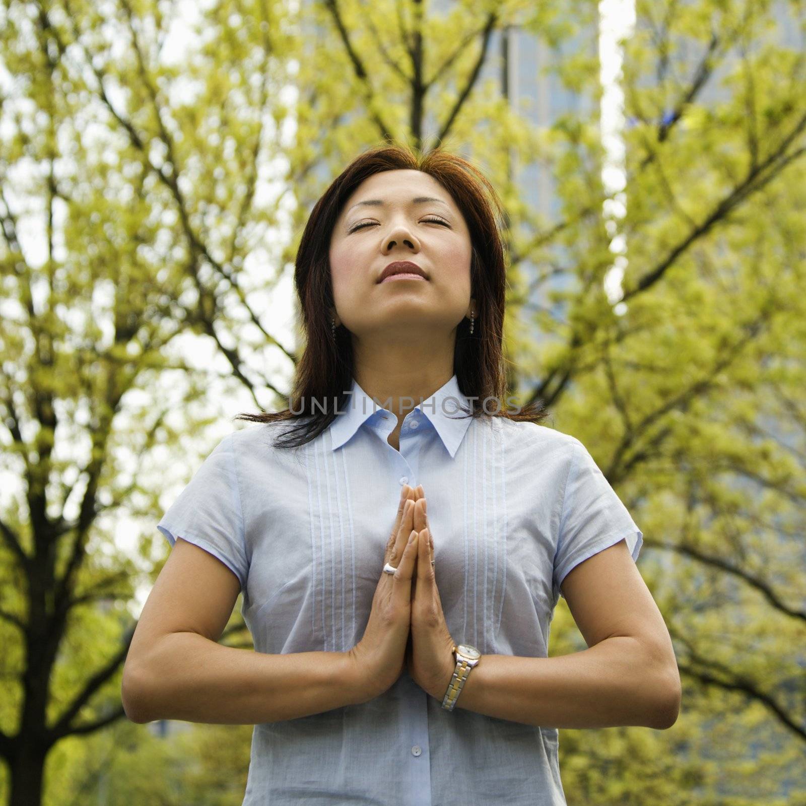 Mid-adult Asian woman standing outside with trees and skyscrapers behind her meditating.