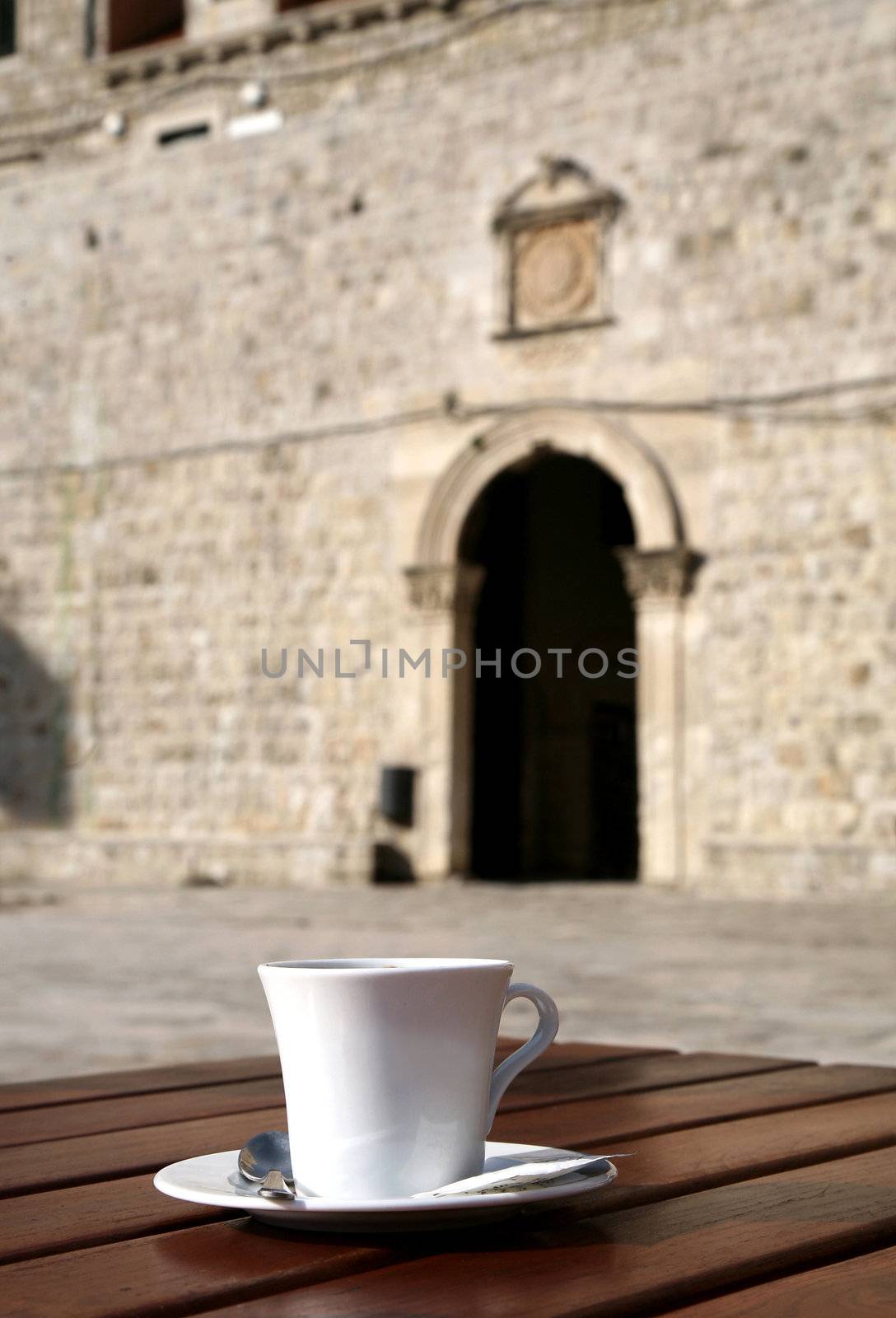 One, white, cup of coffee on table in old harbor in Dubrovnik – Stari Grad. In background-  the gate and curtain walls of the Old Town.