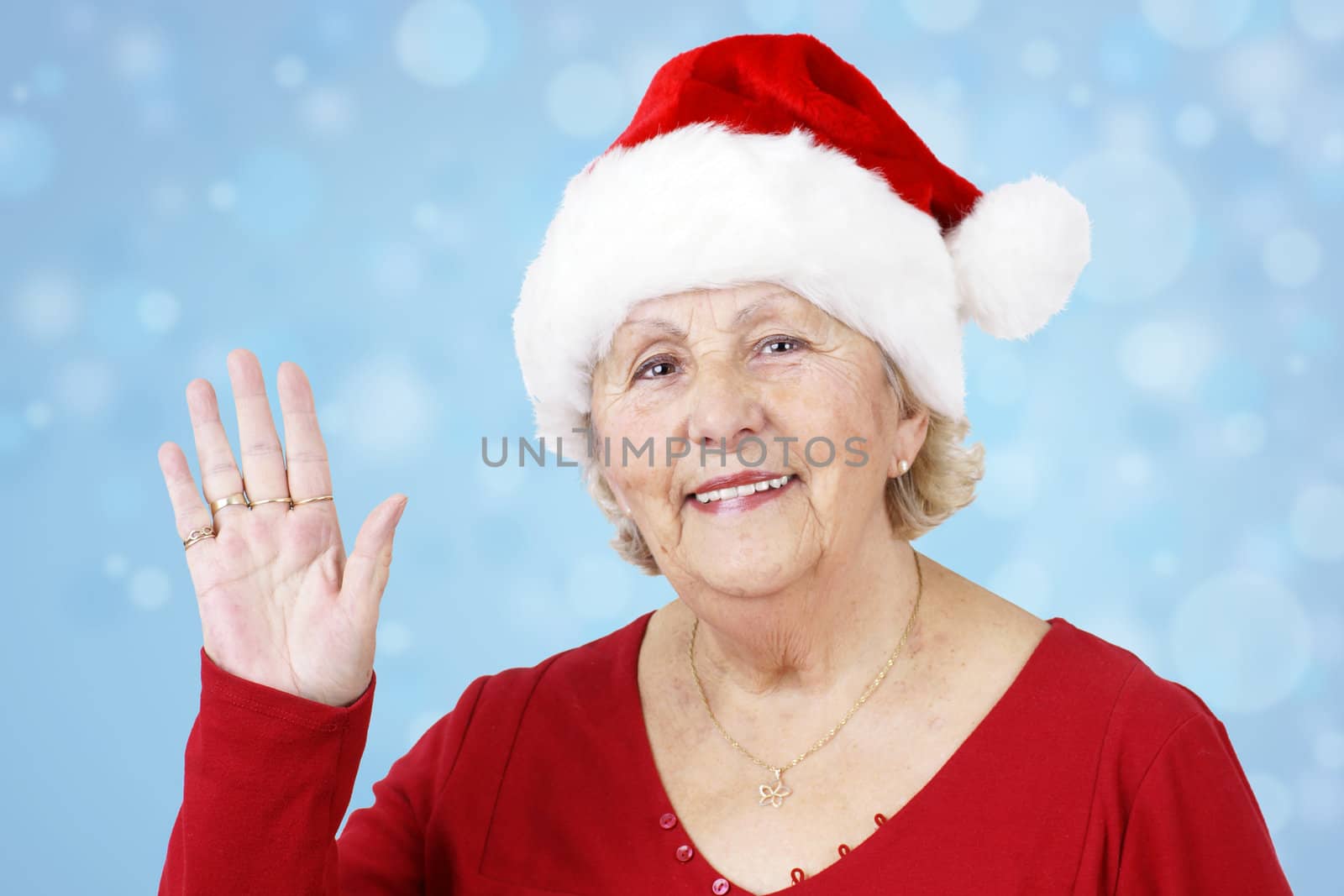 Grand-mother sending her love for Christmas by waving her hand while wearing Santa Claus hat over winter blue background