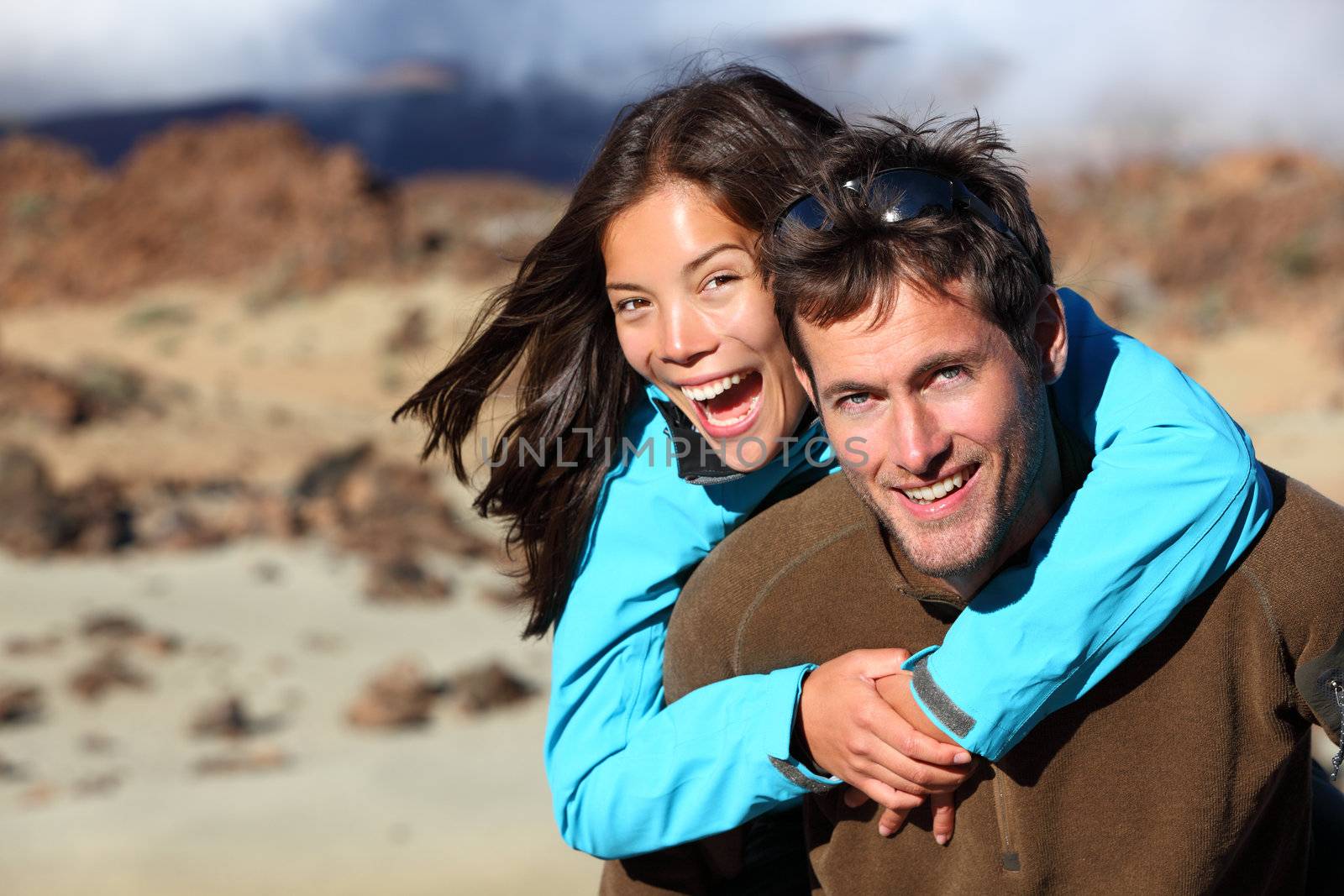 Happy young couple smiling outdoors piggybacking during hiking travel. Young Asian / Caucasian hikers couple cheerful on volcano Teide, Tenerife, Canary Islands.