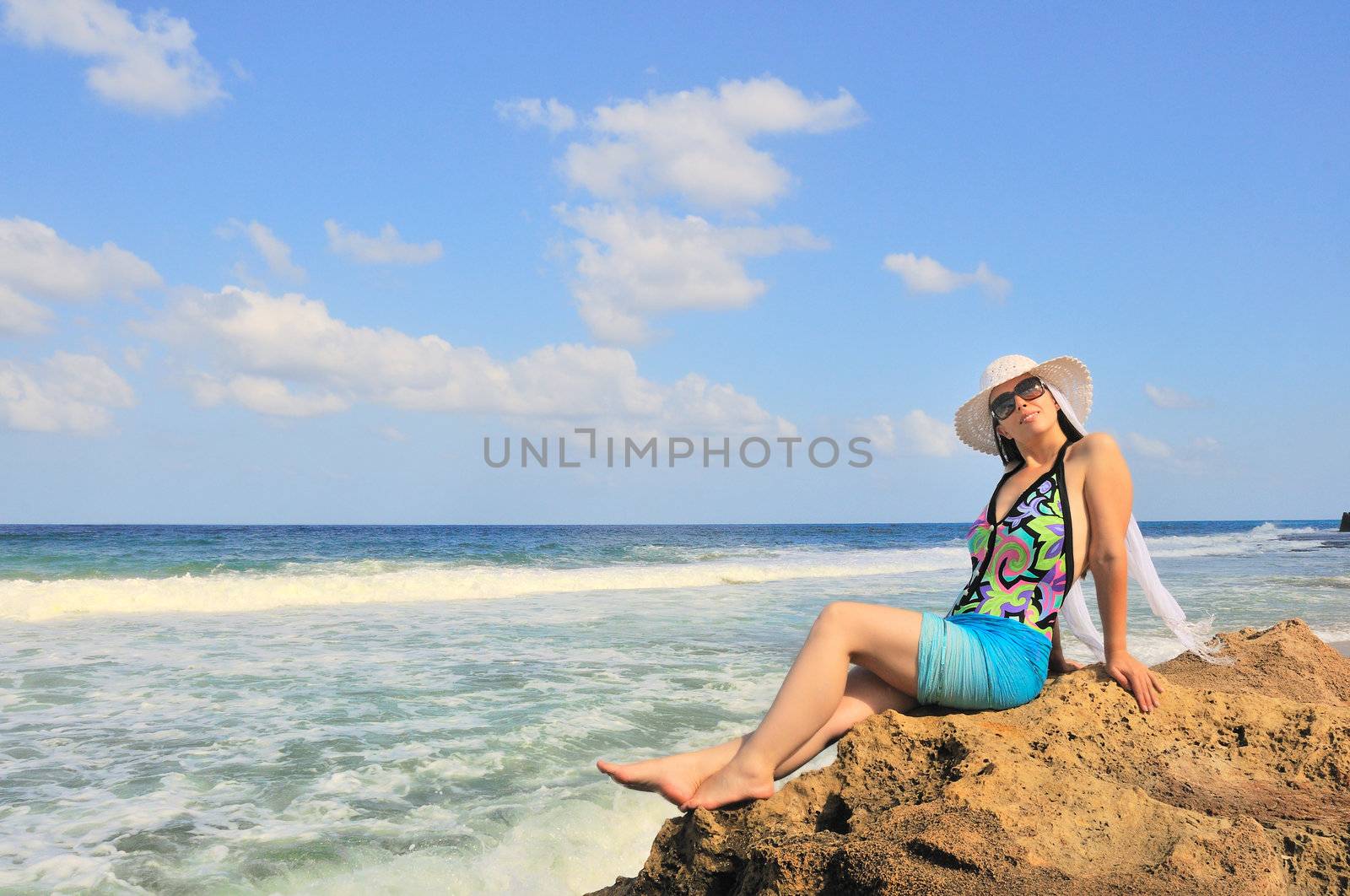 The girl in the white hat sits on a cliff near the sea and posing for a photograph