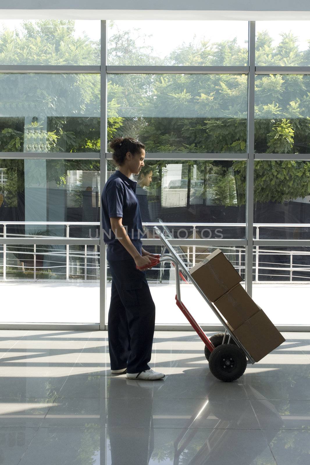 Side view of delivery woman in uniform pushing stack of cardboard boxes on dolly
