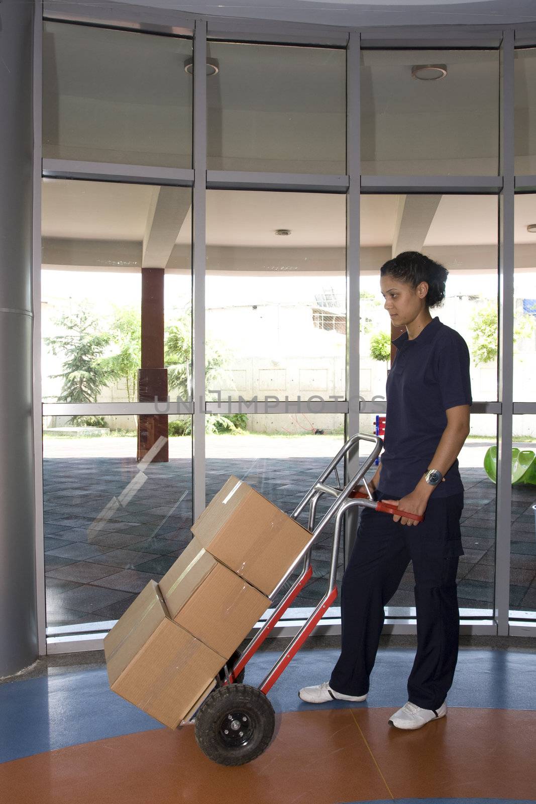 Side view of delivery woman in uniform pushing stack of cardboard boxes on dolly