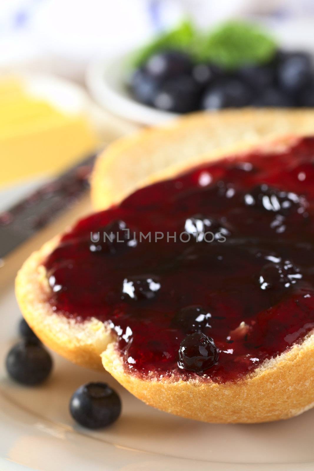 Blueberry jam on half a bun with butter and fresh blueberries in the back (Selective Focus, Focus on the front of the bun and jam)
