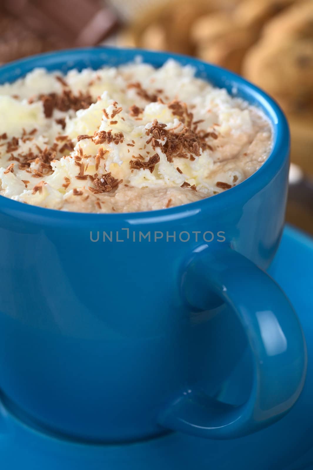 Hot chocolate with whipped cream and chocolate shavings in blue cup with cookies and chocolate in the back (Selective Focus, Focus on the top of the cream)