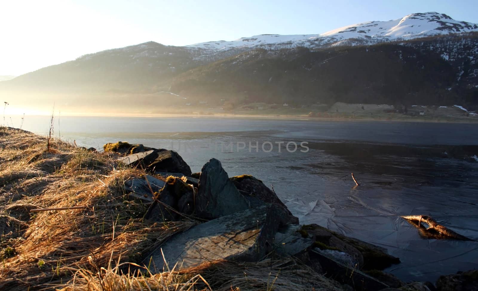 Frosty Morning Norwegian fjord in the foreground illuminated by the sun the grass in the background - snow-capped mountains in the center - water, covered with a crust of ice