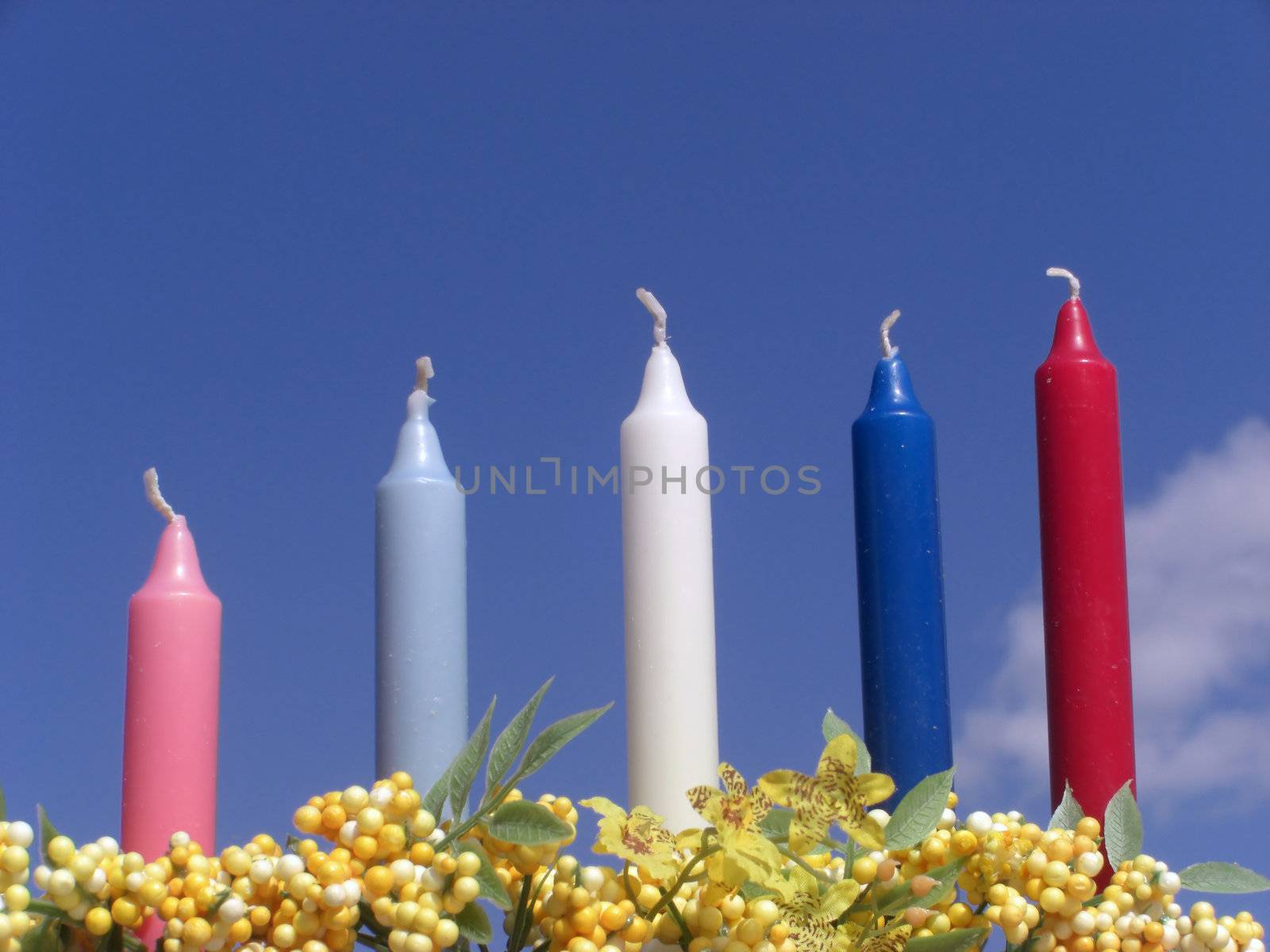 A set of five colorful candles are set in between yellow flowers and against a blue sky.