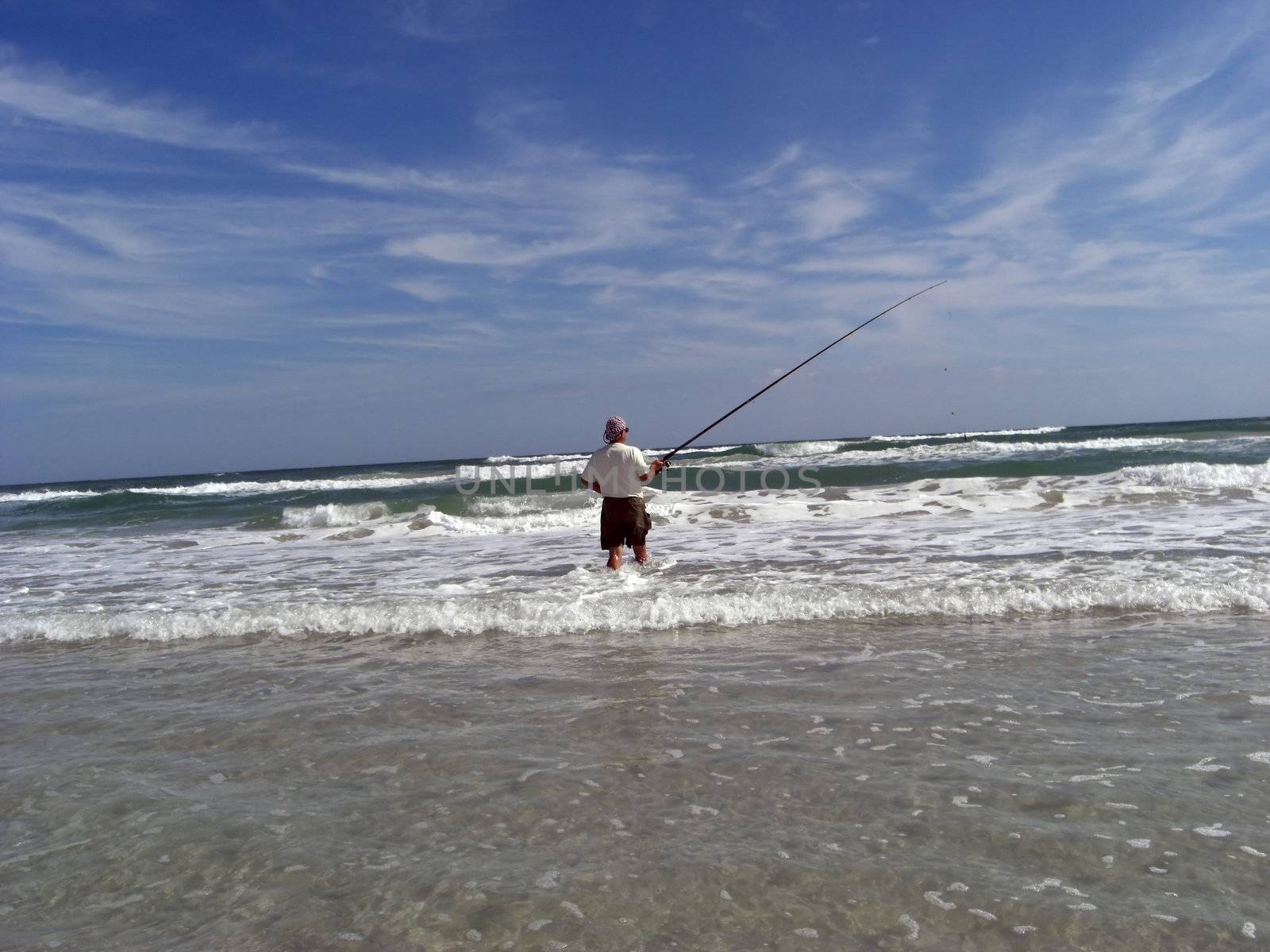 A man is holding a long fishing rod and is fishing for some Bass in a rough ocean.