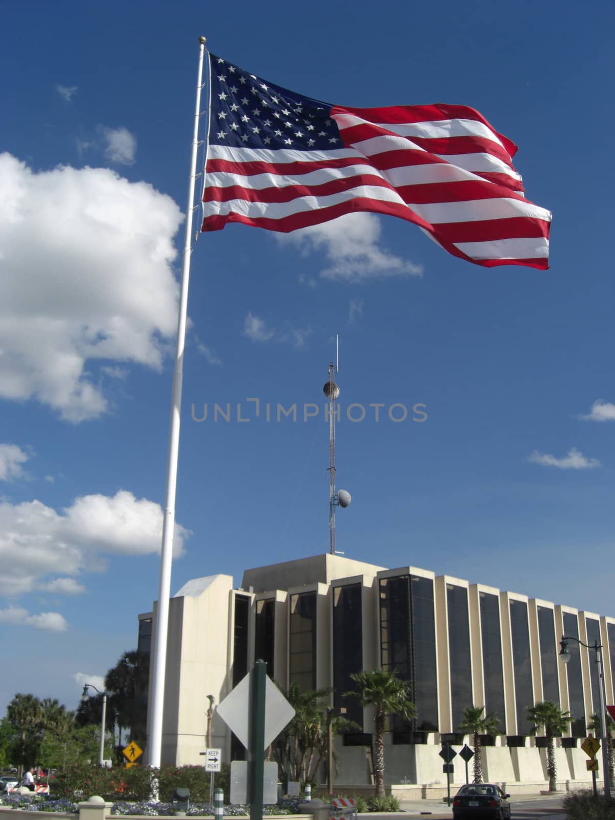 A flag is standing way above the ground and is towering over a building with a satellite on the roof.