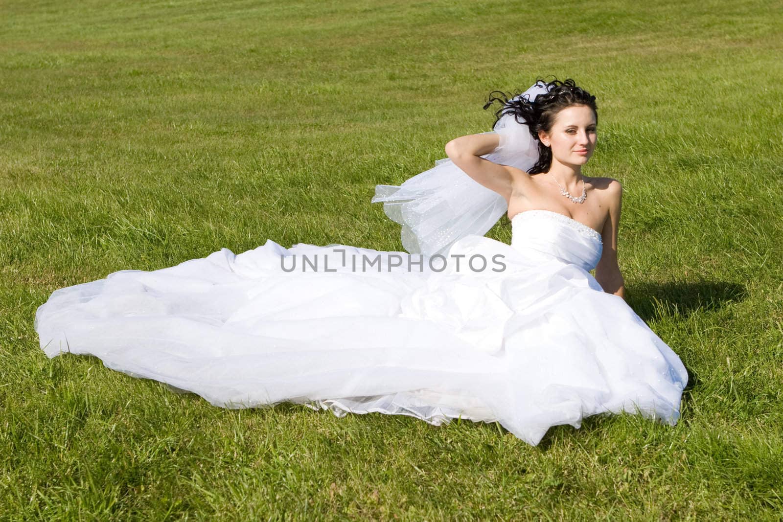 a bride in a beautiful long white dress sits on the green grass and smiles