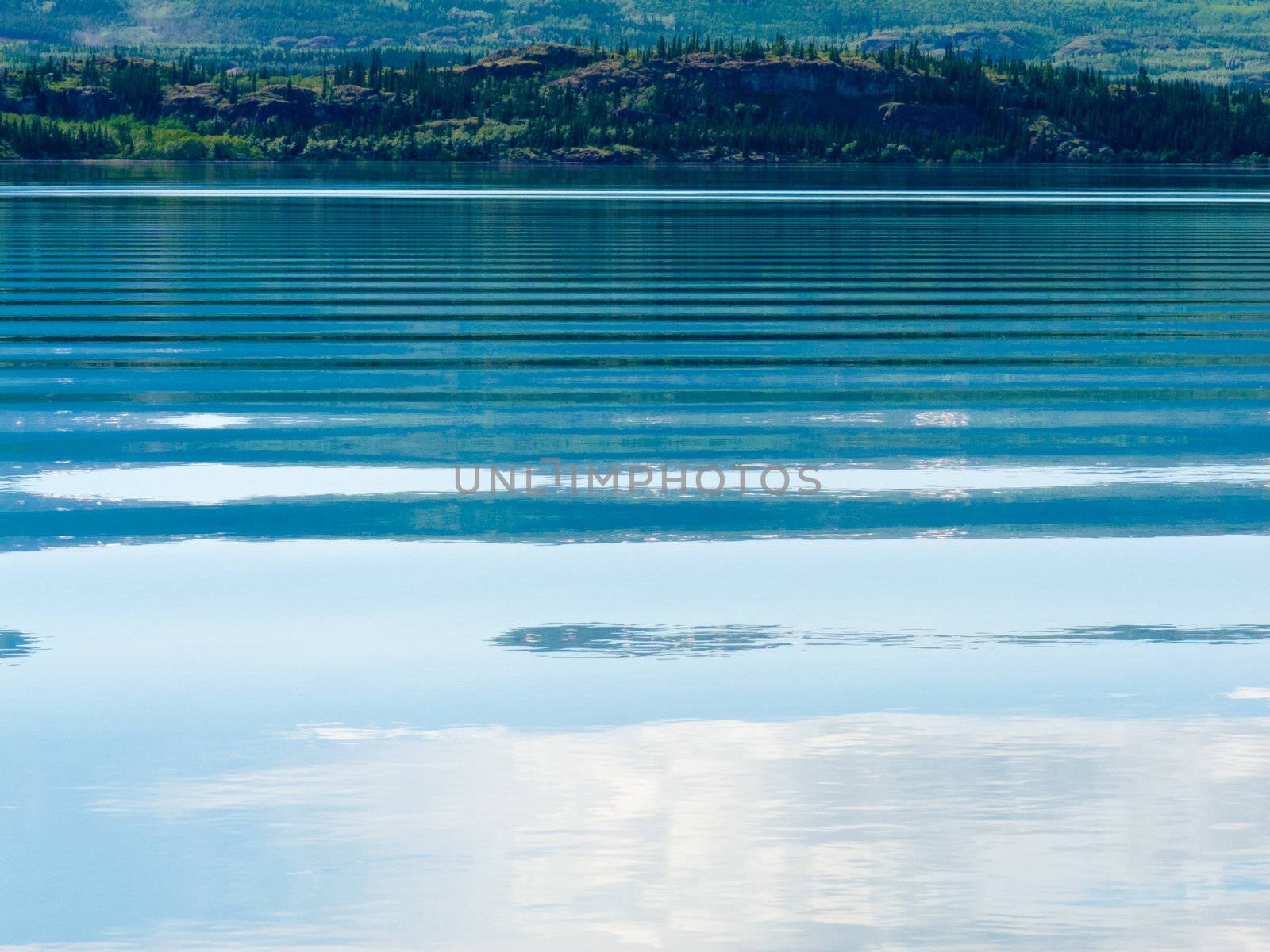 Distant shoreline, clouds and blue sky mirrored on calm lake surface with distortions of some ripples.