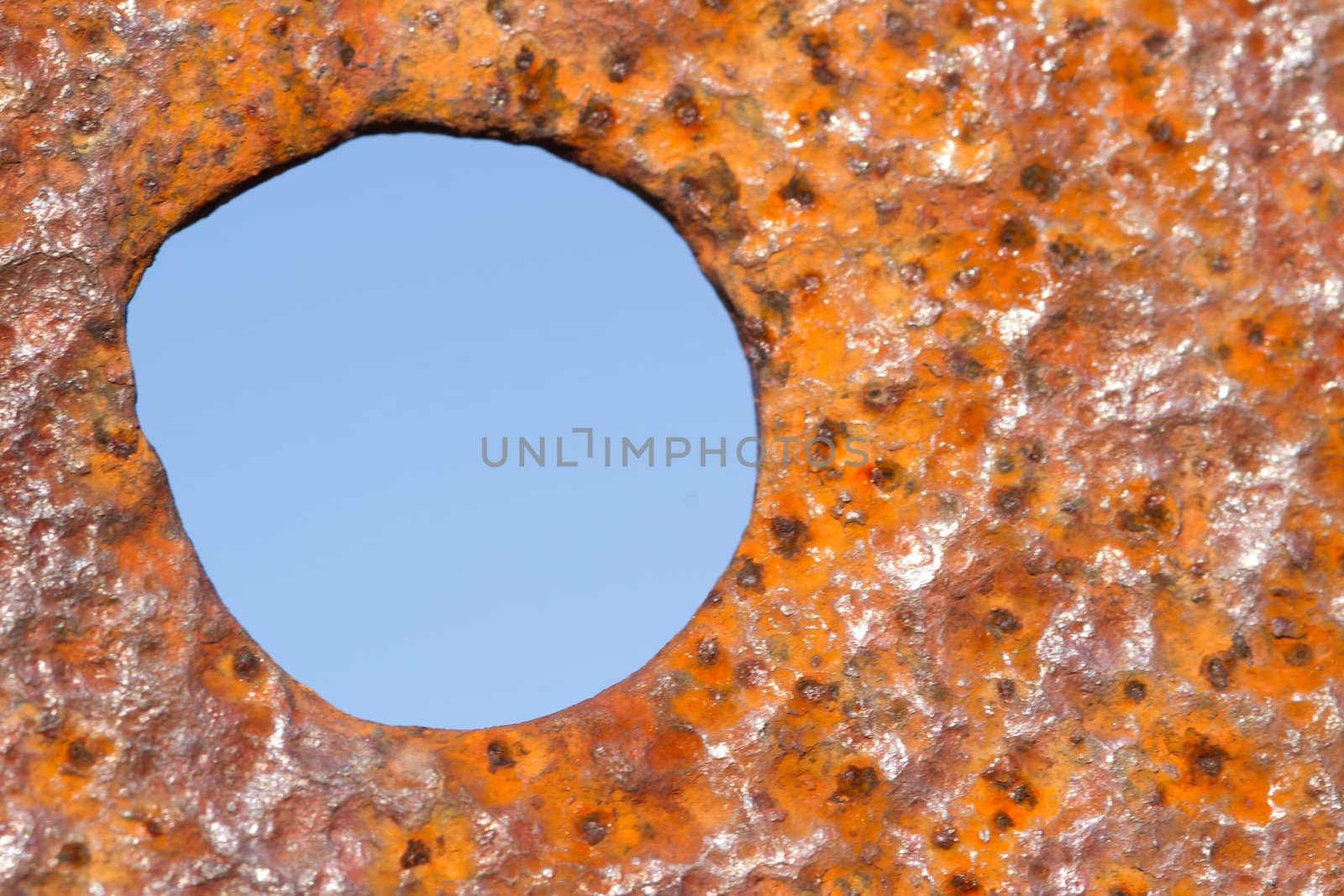 Background of rust, coloured red, orange and yellow with an irregular circle, hole with blue sky beyond.