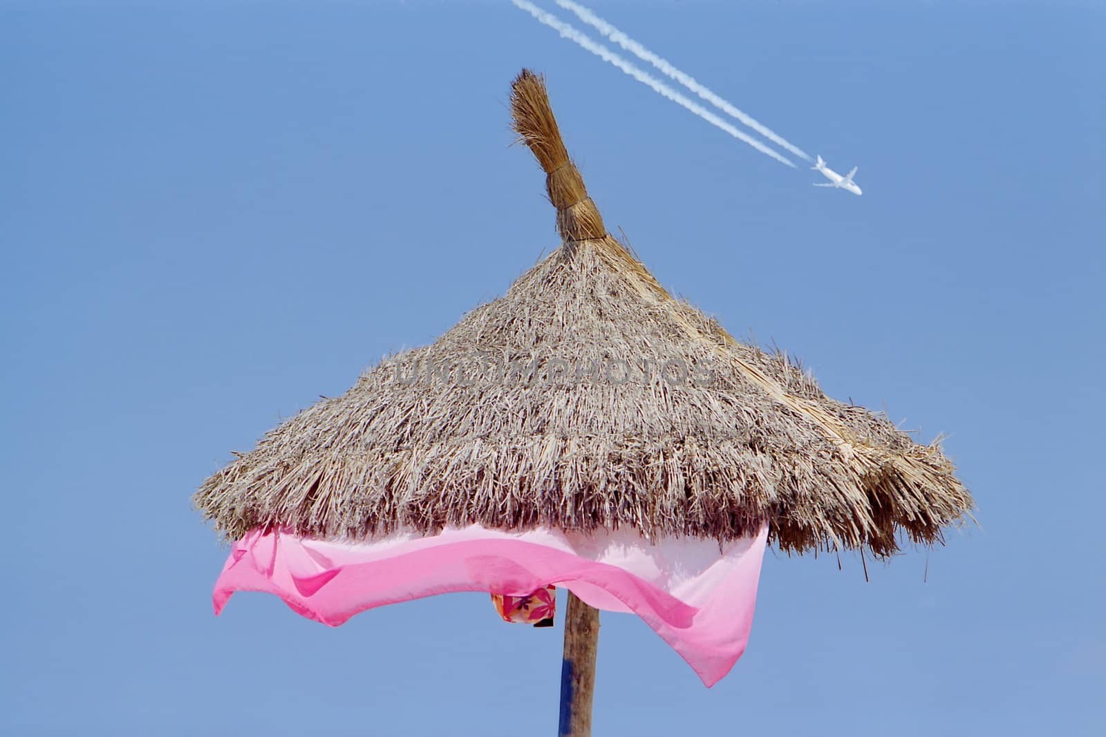 A sun shade of straw against a clear blue sky with an airplane painting white trails, suggesting the consept of summer tourism.