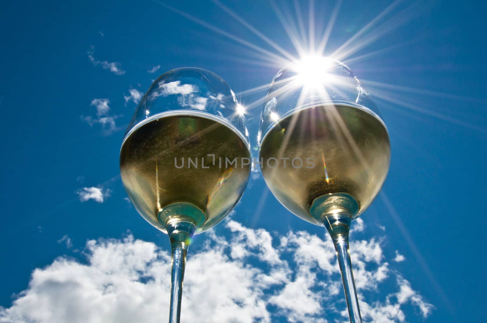 looking up at a closeup of two glasses of white wine & a sunburst through one of them & bright blue sky & clouds in the background.
