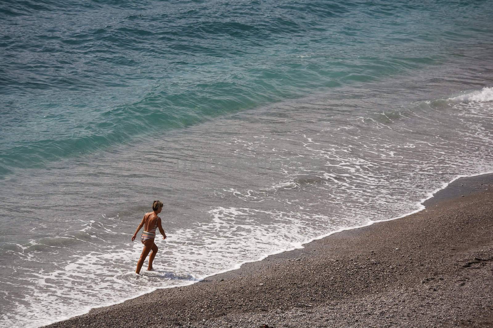 Mature lady walking along the coast on an Italian beach. Space for text.