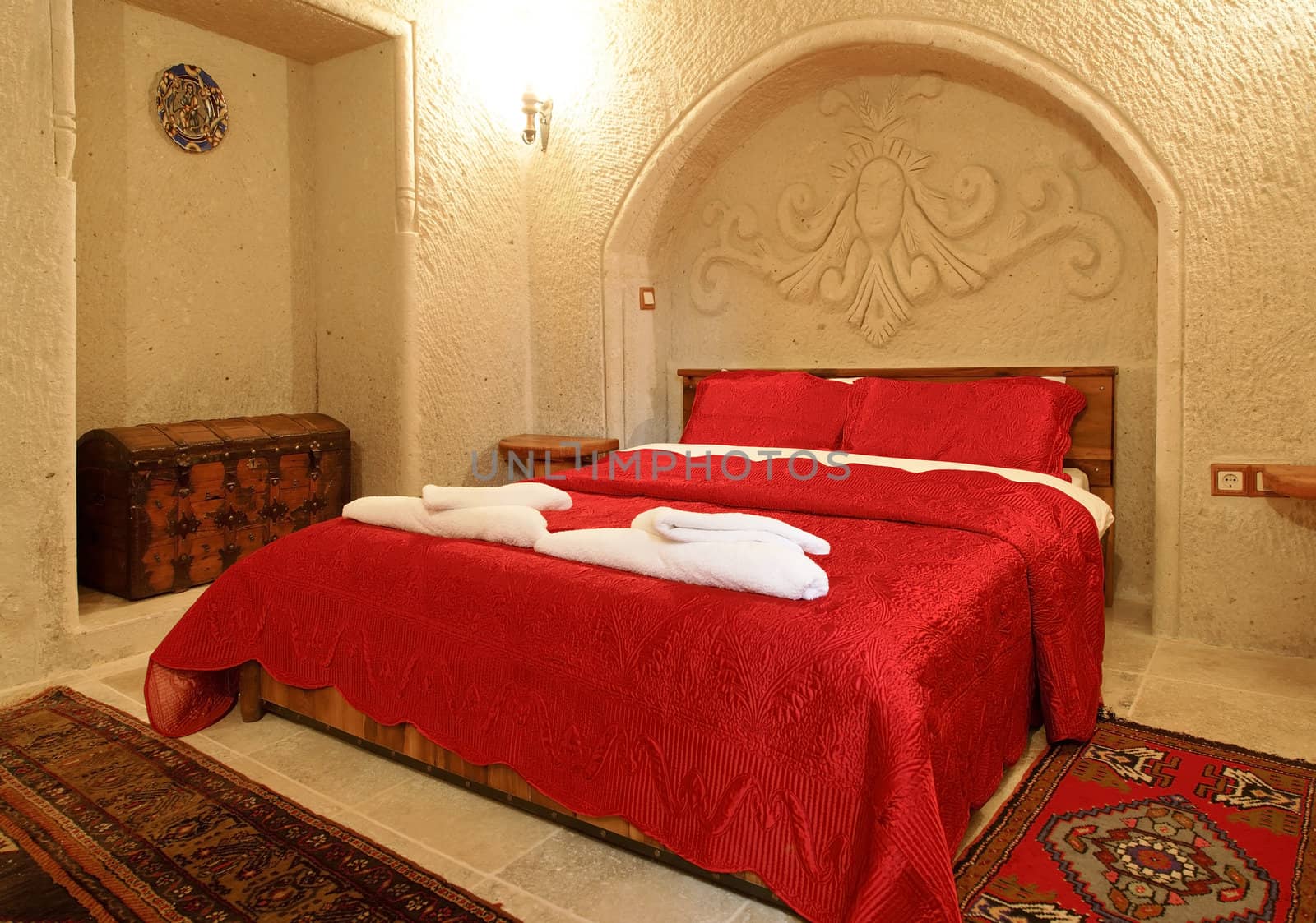 interior architectural detail of bedroom with alcove and archway with detailing, bed cover in embroidered bright red bedspread and pillow cases, treasure chest shipping trunk, woven mats and wall light. Landscape with crop area and copy space.