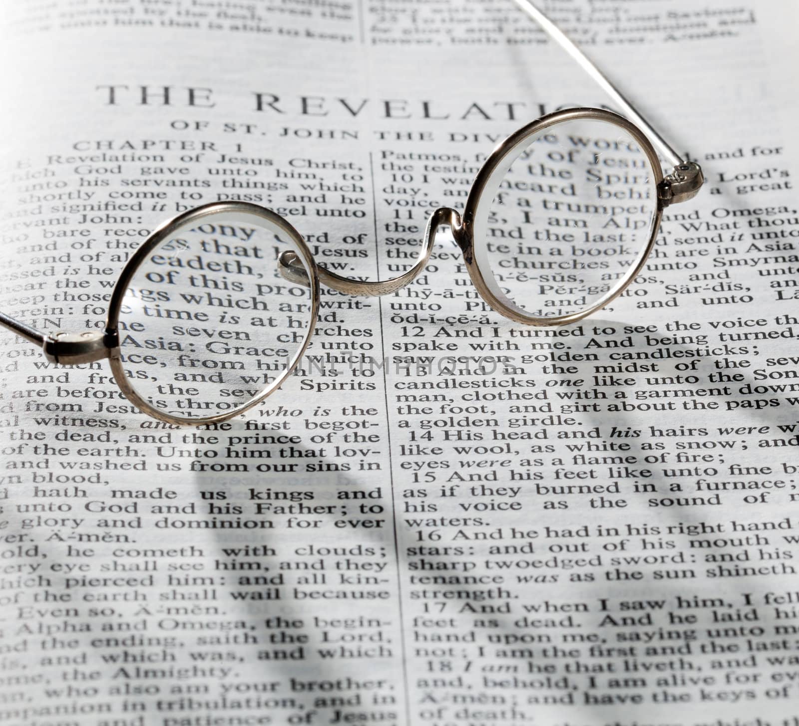 Old fashioned round reading glasses laying on a page from the bible on the revelation with strong shadow and out of focus areas